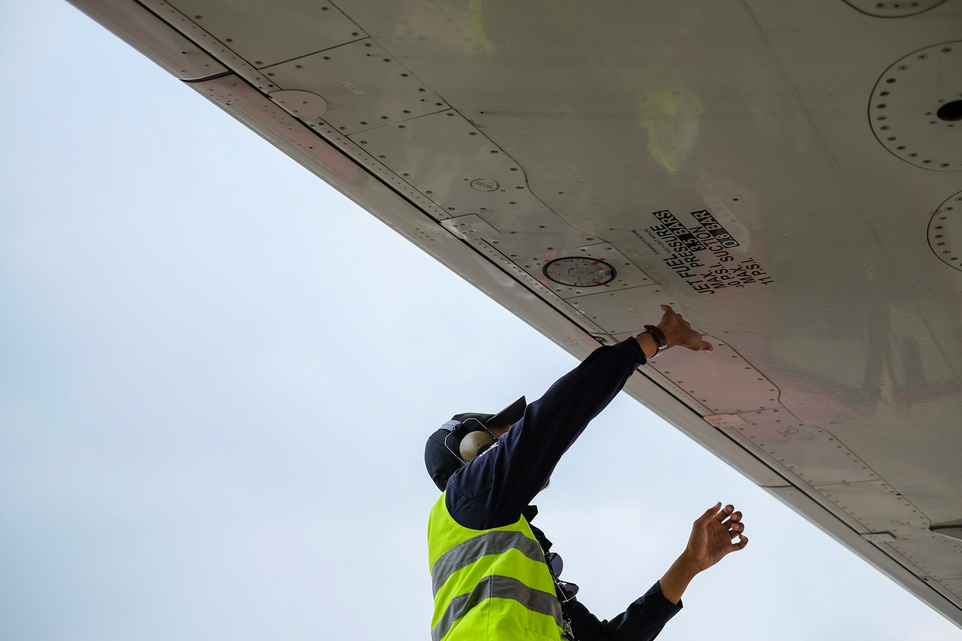 En flytekniker refueller et fly. Mann med refleksvest sett nedenfra under en flyvinge.